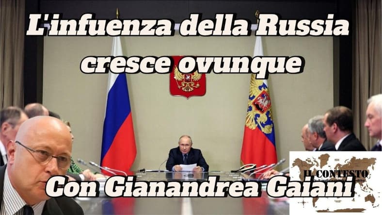 L’influenza della Russia cresce ovunque Un uomo è seduto a un lungo tavolo con dei funzionari, l'influenza della Russia è accentuata dalle bandiere e dagli emblemi sulla parete alle sue spalle.